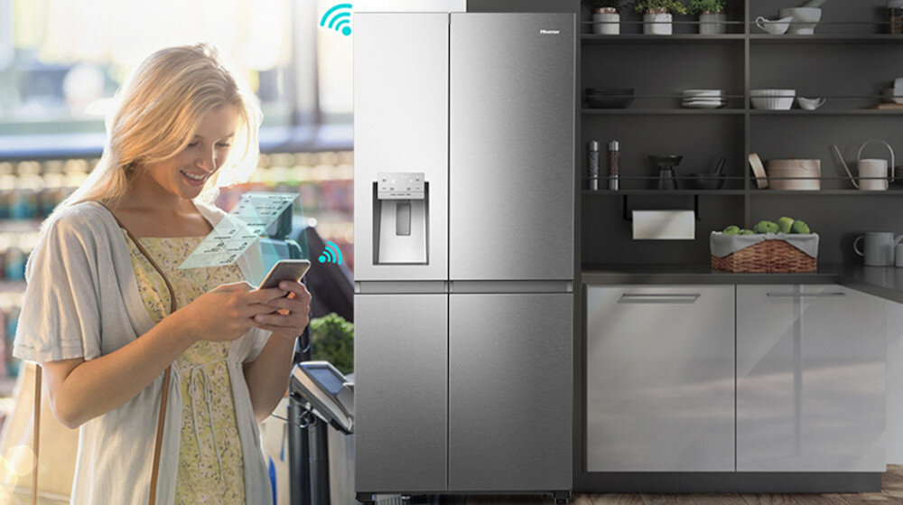 Na obrazie widoczna jest lodówka w kolorze inox z podajnikiem wody i kostkarką, obok której stoi osoba używająca smartfona. W tle znajduje się kuchnia z półkami i szafkami, na których są różne naczynia i kosz z owocami.