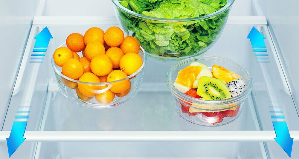 W lodówce znajdują się trzy miski: jedna z sałatą, druga z pomarańczowymi owocami, a trzecia z pokrojonymi owocami, w tym kiwi i pomarańczą. Na półce widoczne są niebieskie strzałki wskazujące przepływ powietrza.