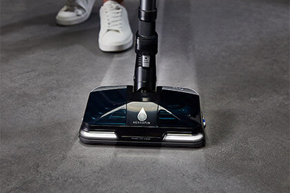 Na obrazie widoczna jest głowica odkurzacza z logo \'AEROSPIN\' na ciemnej powierzchni, znajdująca się na podłodze. W tle widać fragment buta osoby obsługującej urządzenie.
