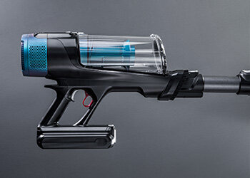 Zdjęcie przedstawia czarny odkurzacz pionowy z przezroczystym pojemnikiem na kurz i niebieskimi elementami. Urządzenie ma ergonomiczny uchwyt i widoczny przycisk w kolorze czerwonym.