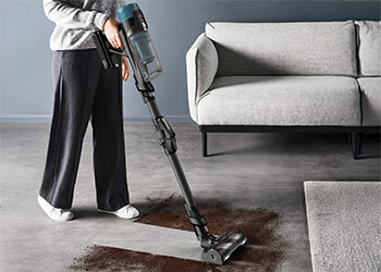 Osoba używa odkurzacza pionowego z przezroczystym pojemnikiem na kurz, czyszcząc brudną powierzchnię podłogi obok szarej kanapy. Odkurzacz ma ciemny kolor i elastyczną rurę.