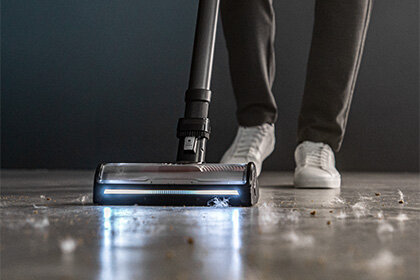 Osoba używa odkurzacza pionowego z podświetlaną szczotką na podłodze z widocznymi drobnymi zanieczyszczeniami. Odkurzacz ma ciemny kolor, a osoba nosi białe buty sportowe.