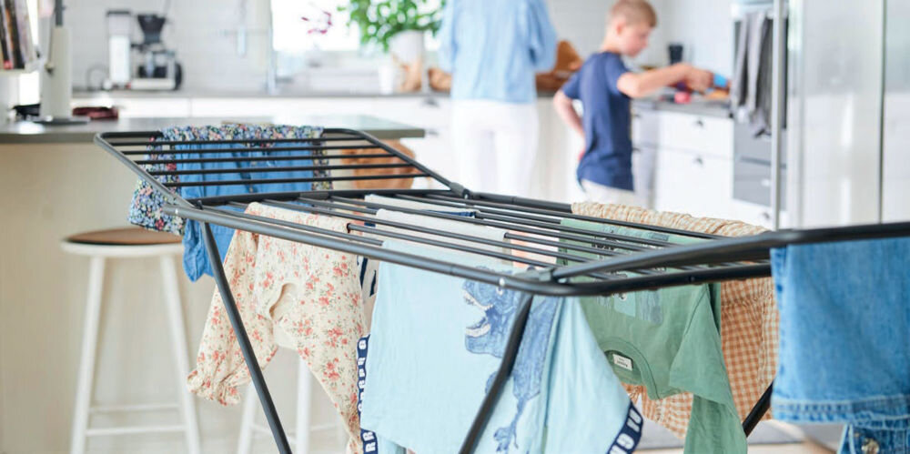 Czarna suszarka na pranie z rozwieszonymi ubraniami stoi w jasnej kuchni. W tle widoczne są osoby zajmujące się codziennymi czynnościami.