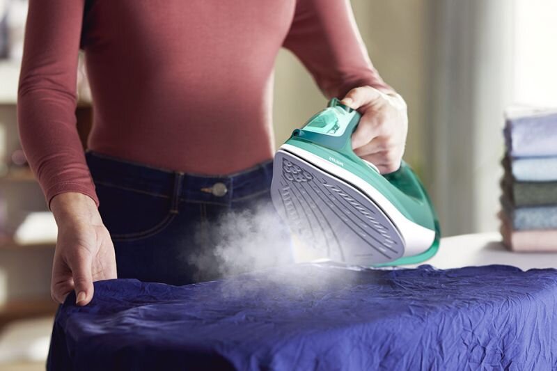 Osoba prasuje niebieski materiał za pomocą zielono-białego żelazka, z którego wydobywa się para. Na żelazku widoczne jest logo \'PHILIPS\'.