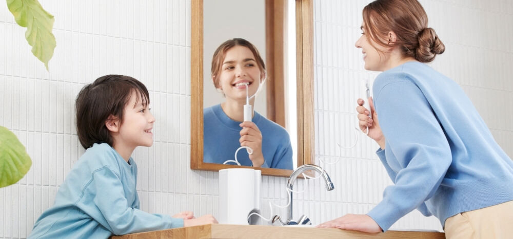 Dwie osoby w łazience używają irygatora przy umywalce. Na blacie stoi biały irygator z widocznym przewodem.