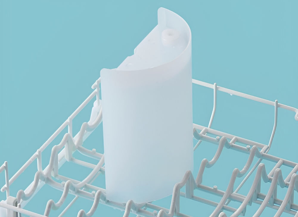 Biały, półprzezroczysty pojemnik umieszczony w koszu zmywarki na tle jasnoniebieskiej ściany. Kosz zmywarki ma metalowe pręty ułożone w regularne wzory.