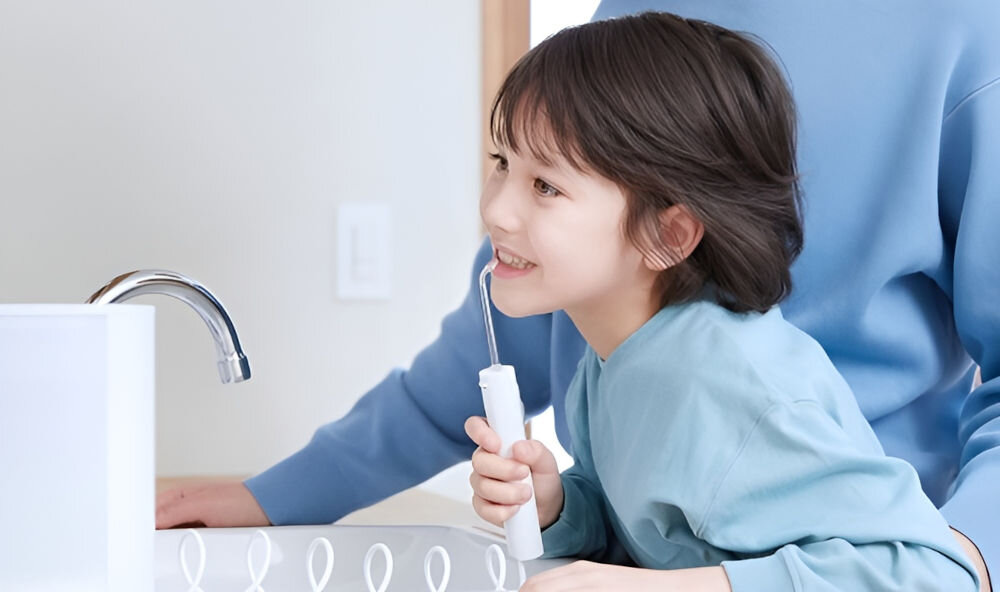 Dziecko trzyma biały irygator przy umywalce, obok stoi dorosła osoba w niebieskiej bluzie. W tle widoczny jest kran i fragment ściany.