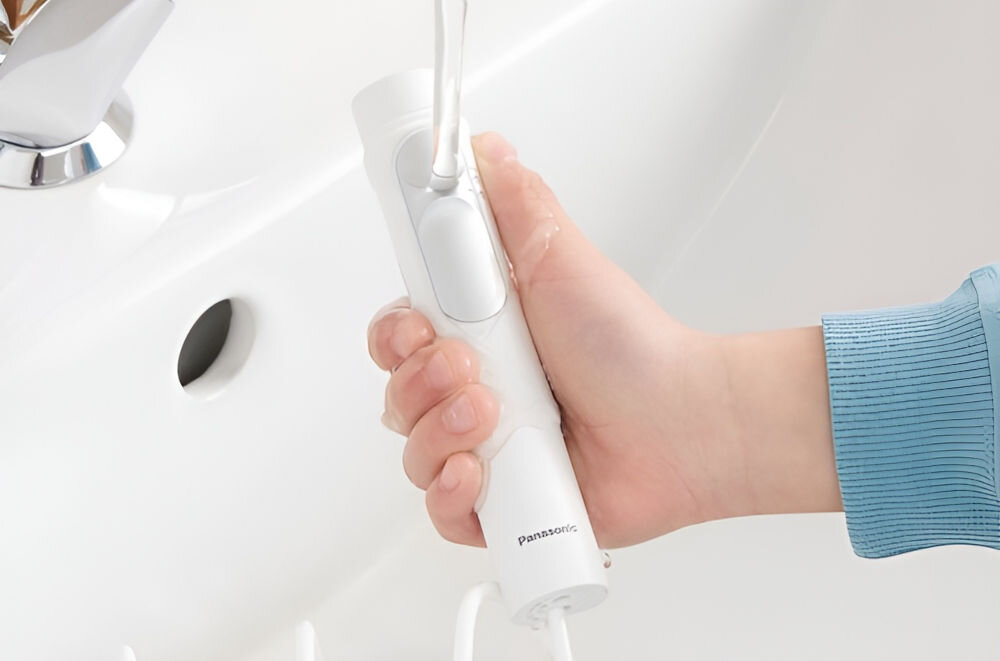 Osoba trzyma biały irygator z widocznym logo \'Panasonic\' nad umywalką. Urządzenie ma smukły kształt i jest podłączone do wężyka.