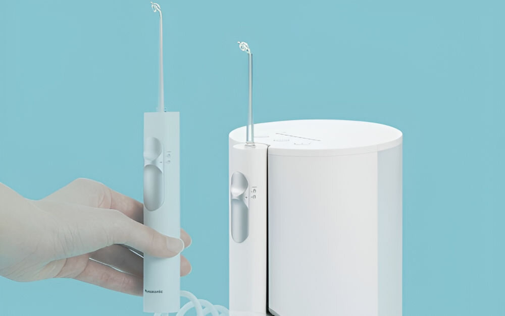 Na obrazie widoczny jest biały irygator z dwoma dyszami, jedna trzymana w dłoni, druga umieszczona w urządzeniu. Na irygatorze widnieje logo \'Panasonic\'.