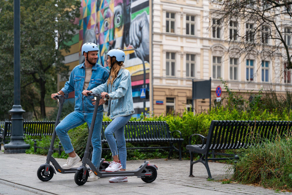 Dwie osoby w kaskach stoją na elektrycznych hulajnogach na tle miejskiego krajobrazu z kolorowym muralem. Hulajnogi są szare, a w tle widać ławki i budynki.