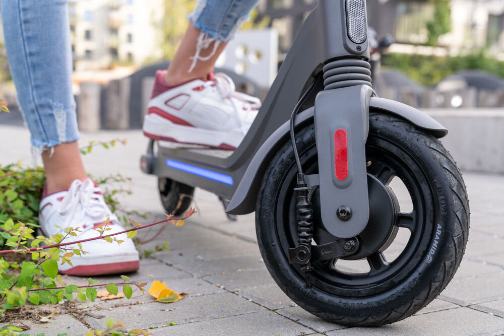 Na zdjęciu widać przednie koło szarej hulajnogi elektrycznej z amortyzacją, obok której stoi osoba w białych butach sportowych. W tle widoczne są elementy miejskiego krajobrazu.
