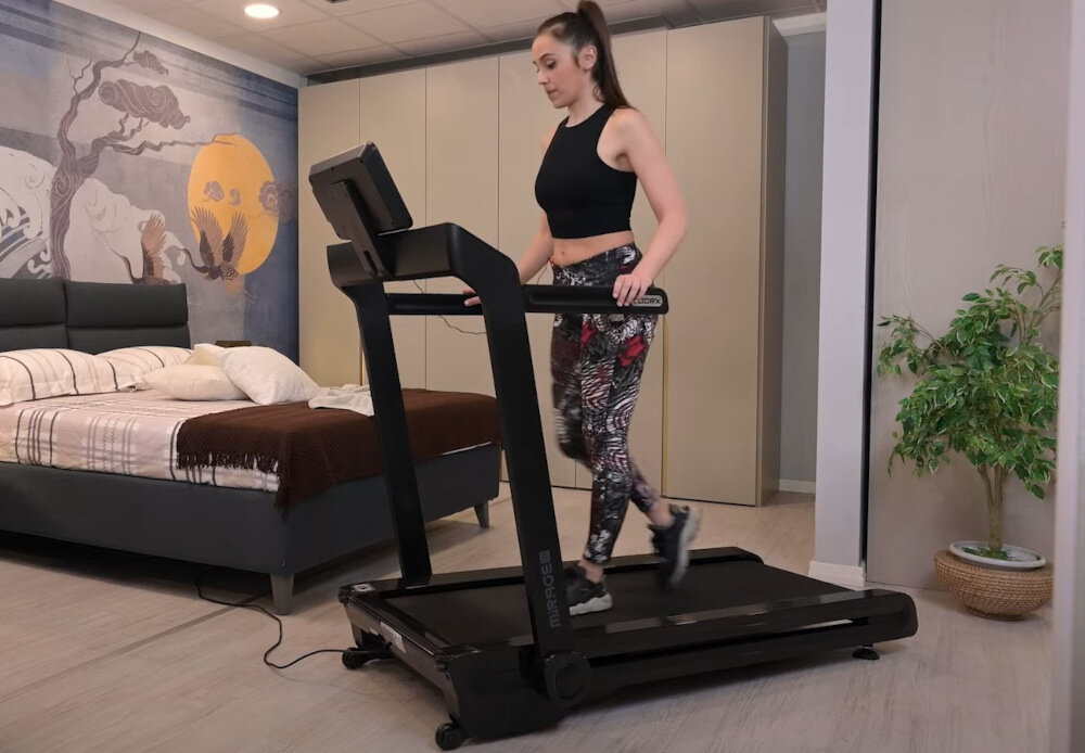 Osoba ćwiczy na bieżni elektrycznej w pokoju z łóżkiem i rośliną w doniczce. Na ścianie widoczna jest dekoracja z motywem krajobrazu.