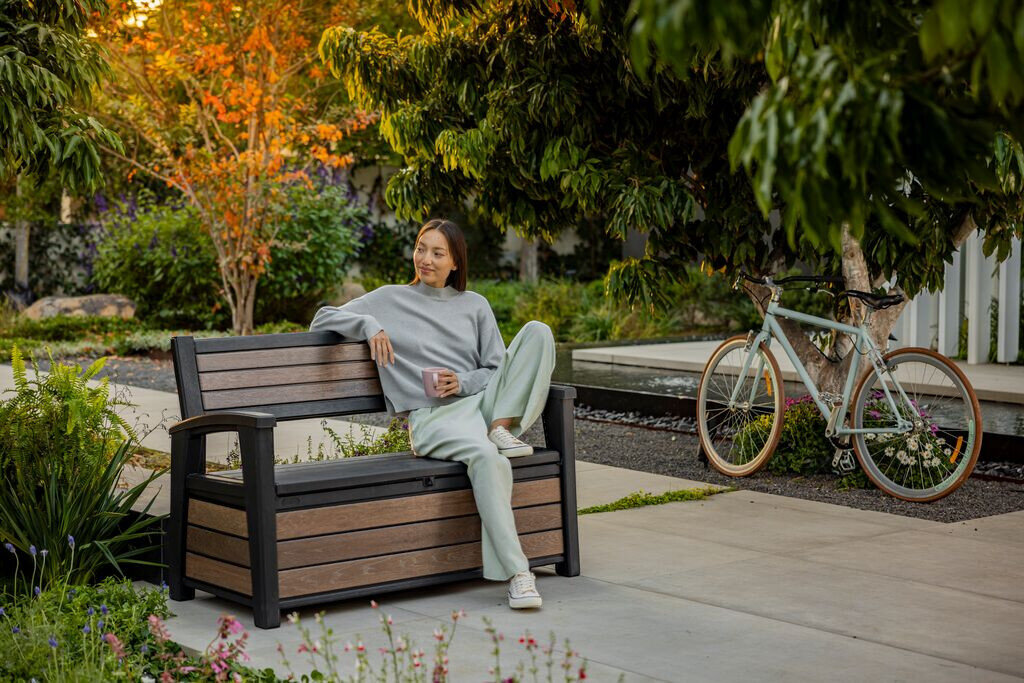 Ławka ogrodowa KETER Deco Garden Bench Brązowy, ławka, ogród, schowek, relaks, Kobieta odpoczywająca na ławce ze schowkiem w ogrodzie obok roweru.