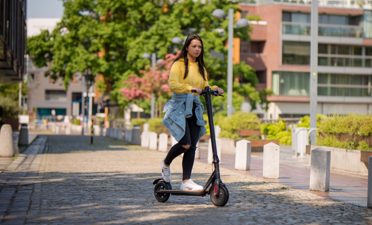 Osoba jedzie na czarnej hulajnodze elektrycznej po brukowanej ścieżce w otoczeniu zieleni i budynków. Hulajnoga ma duże koła i prostą kierownicę.