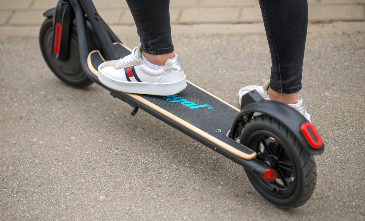 Osoba stoi na czarnej hulajnodze elektrycznej z niebieskim napisem \'frugal\' na podeście. Hulajnoga ma czerwone elementy przy kołach i znajduje się na chodniku.