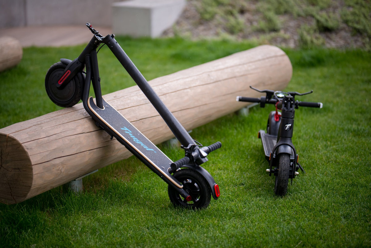 Dwie czarne hulajnogi elektryczne stoją na trawie obok drewnianego pnia. Na jednej z nich widoczny jest napis \'Frugal\'.