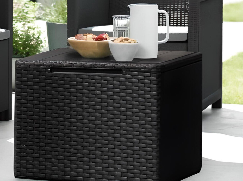 Na zdjęciu widoczny jest ciemny, pleciony stolik ogrodowy z tacą na górze, na której znajdują się dzbanek, szklanka oraz miski z przekąskami. W tle widać fragmenty innych mebli ogrodowych oraz zieleń.