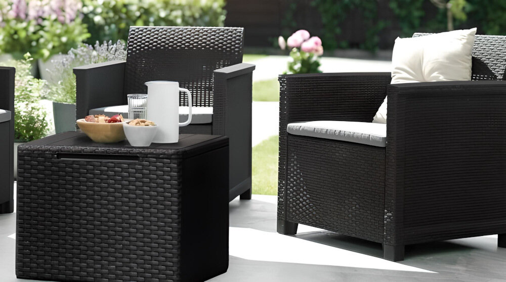 Na obrazie widoczny jest zestaw mebli ogrodowych składający się z dwóch foteli i stolika w kolorze grafitowo-jasnoszarym. Na stoliku znajdują się dzbanek i miski z przekąskami, a w tle widać rośliny ogrodowe.