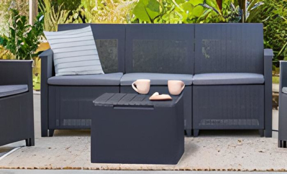 Zestaw mebli ogrodowych w kolorze grafitowo-jasnoszarym, składający się z sofy z poduszkami, stolika oraz dwóch kubków i talerzyka na blacie. W tle widoczna jest roślinność ogrodowa.