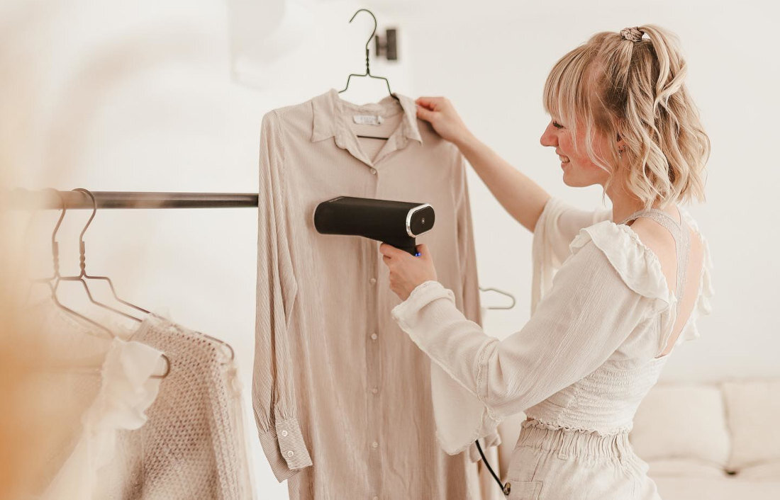 Osoba używa parownicy do ubrań na zawieszonej koszuli. W tle widać jasne wnętrze z wieszakiem na ubrania.