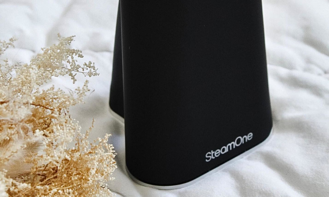 Czarna podstawa urządzenia z widocznym logo \'SteamOne\' na białym tle. Obok znajduje się sucha roślinność na białej powierzchni.