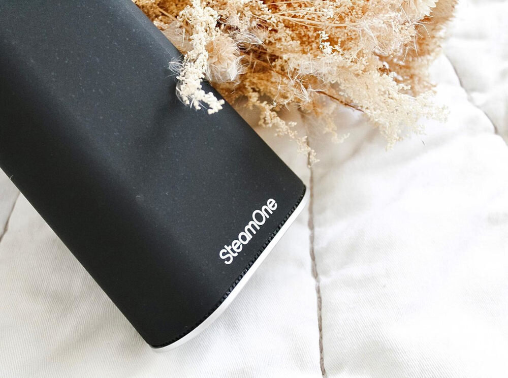 Czarny element urządzenia z widocznym logo \'SteamOne\' na jasnym tle. Obok znajduje się sucha roślinność o jasnym kolorze.