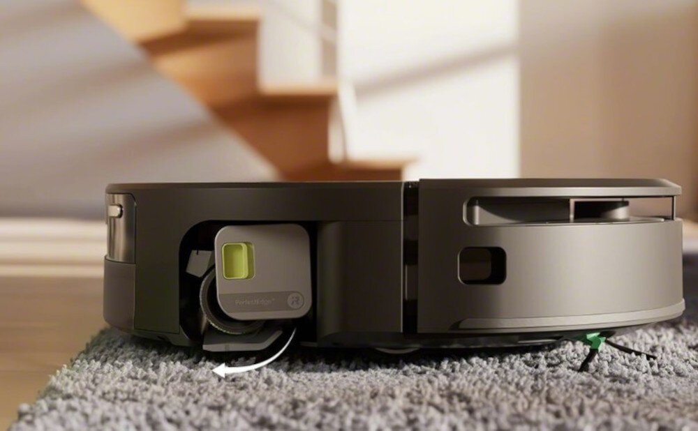 Na obrazie widoczny jest czarny robot sprzątający na dywanie, z otwartą klapką ukazującą wnętrze urządzenia. W tle znajdują się schody i rozmyte elementy wnętrza.