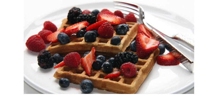 Na białym talerzu znajdują się dwa gofry udekorowane świeżymi owocami, takimi jak truskawki, maliny, jeżyny i borówki. Obok leżą sztućce: widelec i nóż.