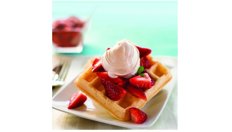 Na talerzu znajduje się gofr z kawałkami truskawek i kleksem bitej śmietany. W tle widoczna jest miska z truskawkami.