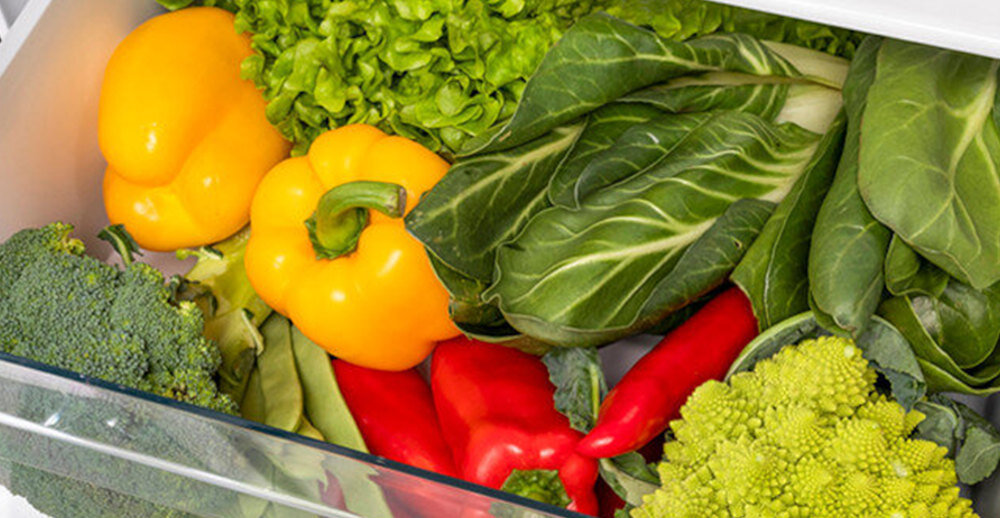 W szufladzie lodówki znajdują się różne warzywa, w tym żółta papryka, czerwona papryka, brokuły, sałata i liście zielone. Warzywa są świeże i kolorowe.
