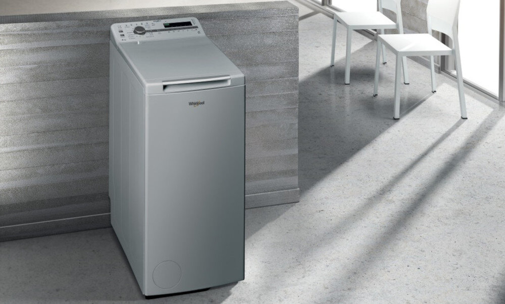 Srebrna pralka ładowana od góry stoi w nowoczesnym wnętrzu. Na panelu sterowania widoczne są przyciski i pokrętło, a na froncie logo Whirlpool.