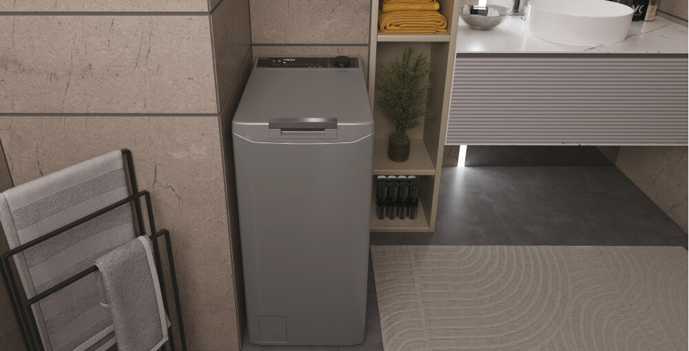 Na obrazie znajduje się szara pralka w nowoczesnej łazience, obok której znajduje się regał z ręcznikami i kosmetykami. Pralka jest umieszczona obok umywalki na szafce.