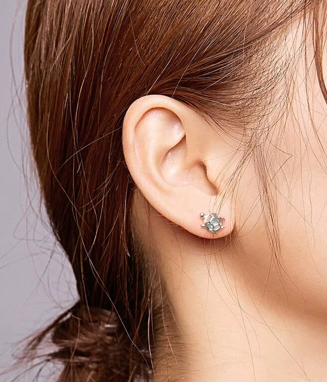 Zdjęcie przedstawia ucho z założonym srebrnym kolczykiem w kształcie żółwia, ozdobionym niebieskimi kamieniami. Włosy są brązowe i związane z tyłu.