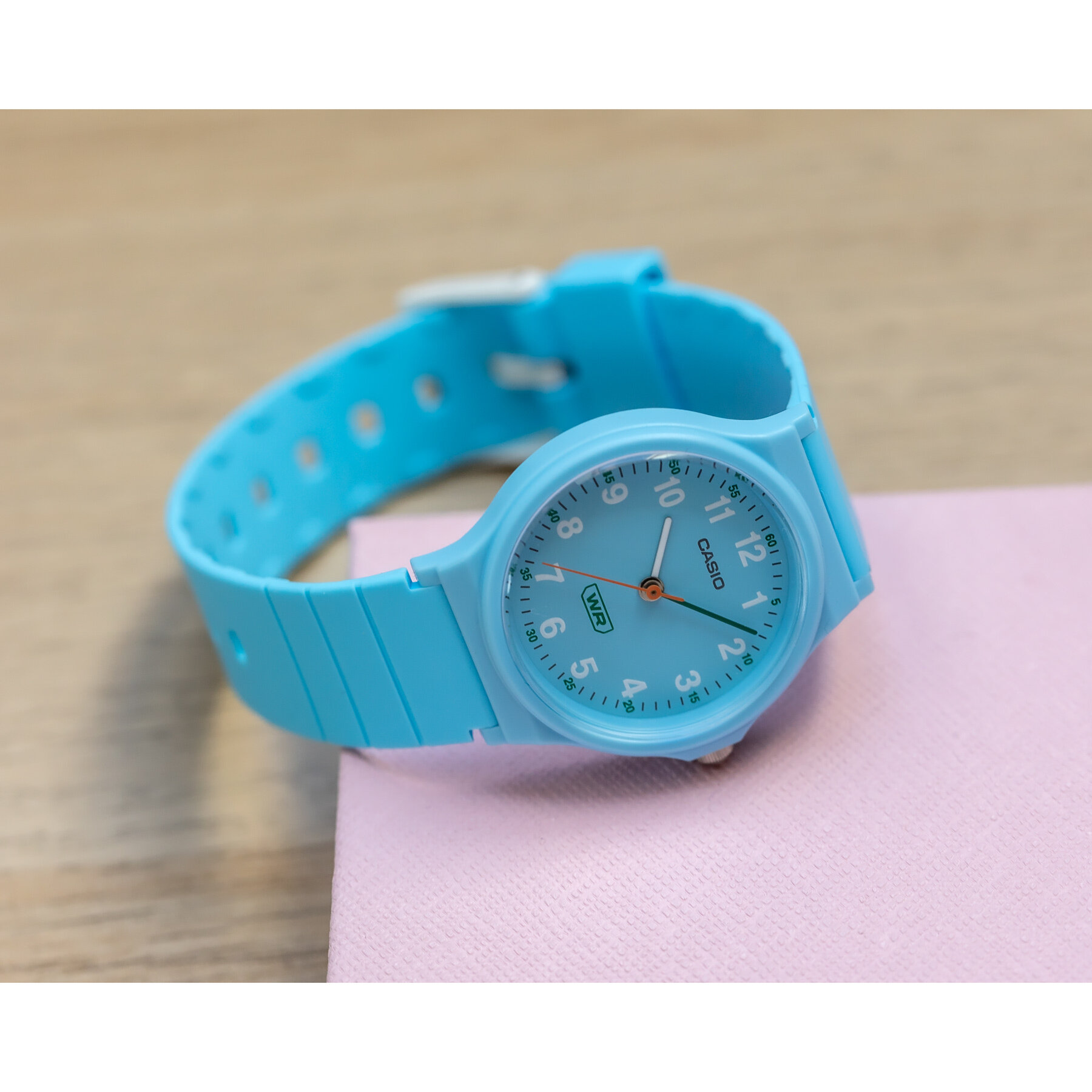 Jasnoniebieski zegarek z okrągłą tarczą, na której widoczne są cyfry arabskie i logo Casio. Zegarek leży na różowej powierzchni.