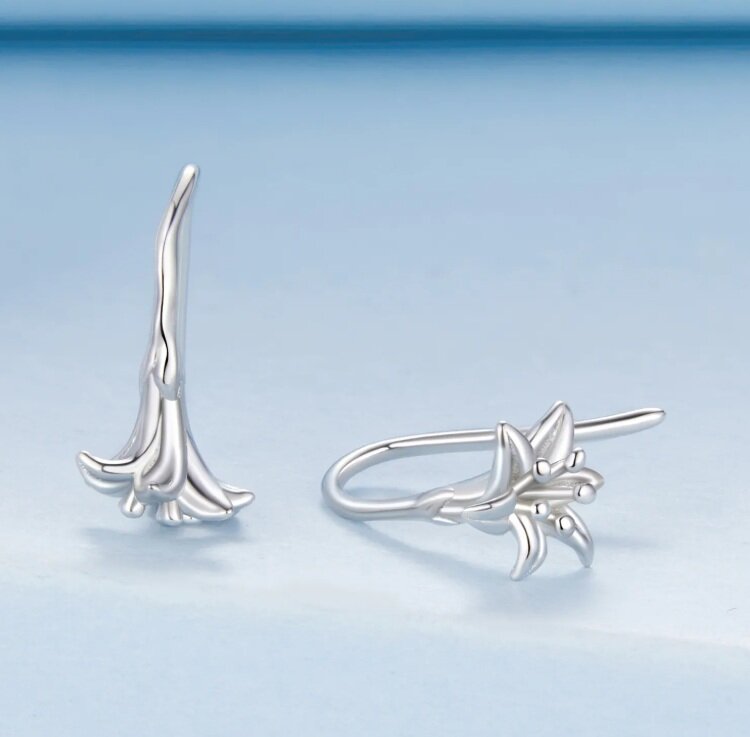 Na obrazie widoczne są srebrne kolczyki w kształcie kwiatu lilii, wykonane z metalu o gładkiej powierzchni. Kolczyki mają delikatny, elegancki design z wyraźnie zaznaczonymi płatkami.
