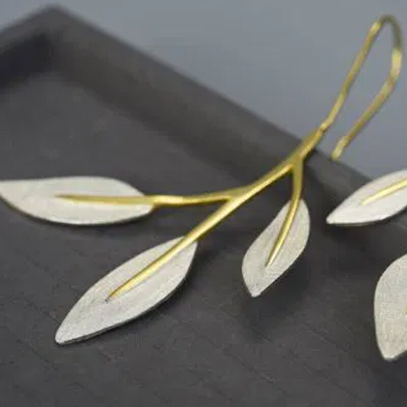 Na obrazie widoczne są kolczyki w kształcie gałązki z liśćmi, wykonane z pozłacanego srebra. Kolczyki leżą na ciemnym tle.
