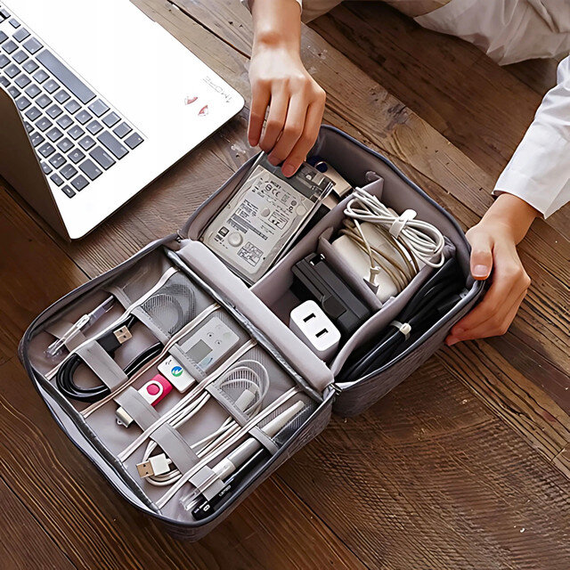 Na drewnianym stole leży otwarty organizer z różnymi kablami, dyskiem zewnętrznym i akcesoriami elektronicznymi. Obok znajduje się laptop, a ręce osoby trzymają organizer.
