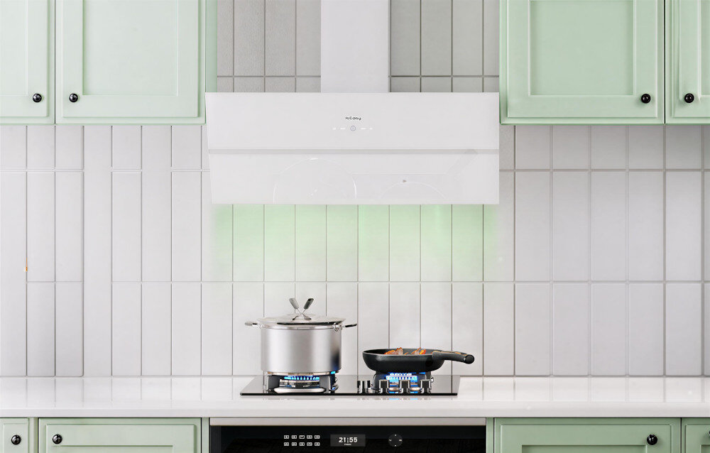 Biały okap kuchenny zamontowany nad kuchenką z garnkiem i patelnią na palnikach. W tle widoczne są jasne płytki ścienne oraz zielone szafki kuchenne.