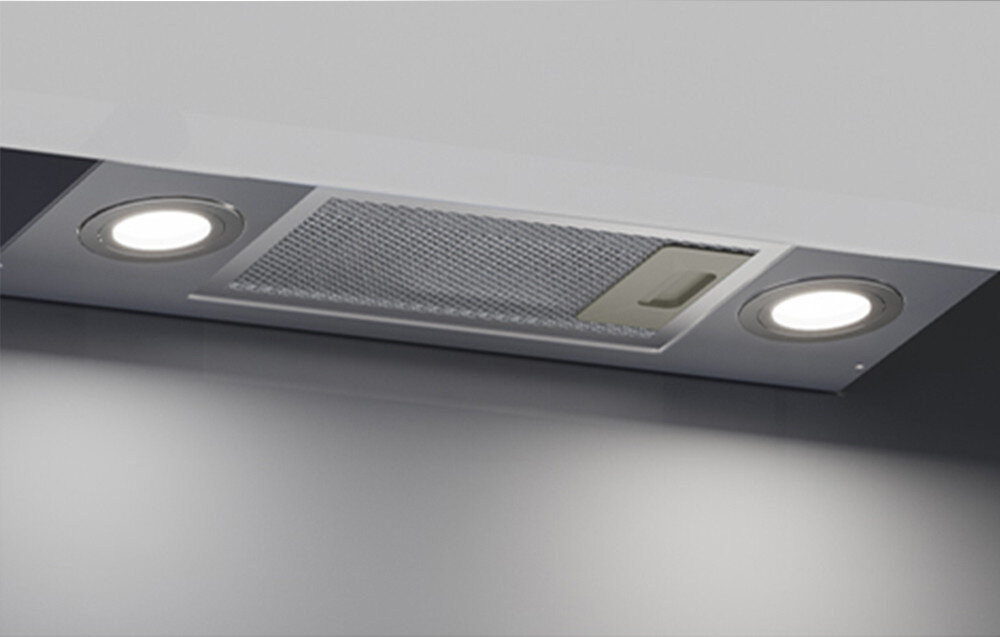 Na obrazie widoczny jest biały okap kuchenny z dwoma okrągłymi światłami po bokach i metalowym filtrem pośrodku. Okap jest zamontowany w suficie lub szafce.