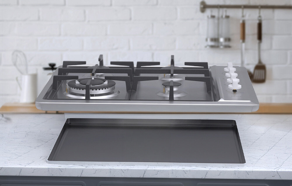 Na obrazie widoczna jest płyta gazowa z czterema palnikami i pokrętłami do regulacji. W tle znajduje się biała ceglana ściana z zawieszonymi przyborami kuchennymi.