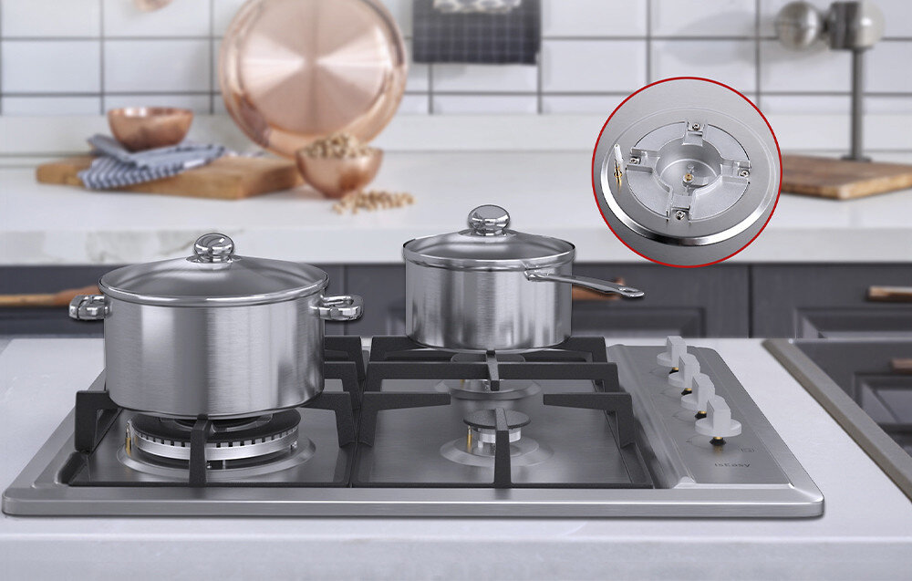 Na obrazie widoczna jest płyta gazowa z czterema palnikami, na której stoją dwa garnki. W prawym górnym rogu znajduje się powiększenie jednego z palników, a w tle widać kuchenne akcesoria.