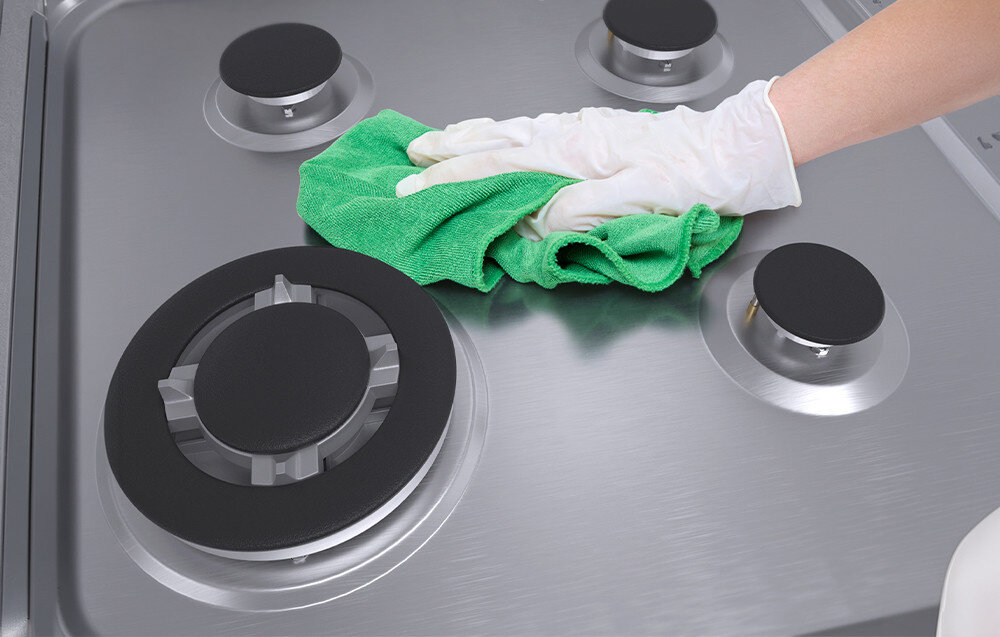 Osoba w rękawiczce czyści zieloną ściereczką płytę gazową z czterema palnikami. Powierzchnia płyty jest metaliczna i błyszcząca.