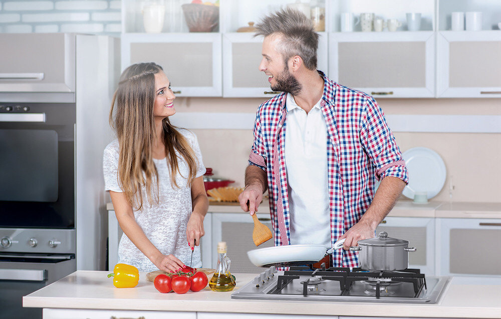 Dwie osoby gotują w kuchni, używając płyty gazowej z garnkiem i patelnią. Na blacie znajdują się pomidory, żółta papryka i butelka z oliwą.