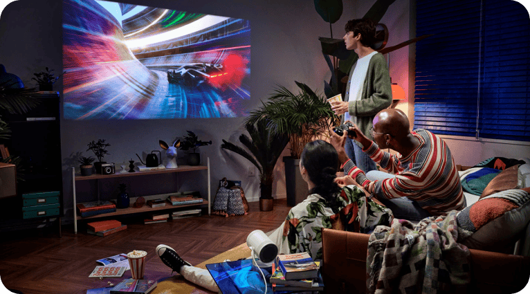 W salonie na ścianie wyświetlany jest dynamiczny obraz wyścigu samochodowego. Kilka osób siedzi na kanapie, oglądając projekcję z przenośnego projektora umieszczonego na stoliku.