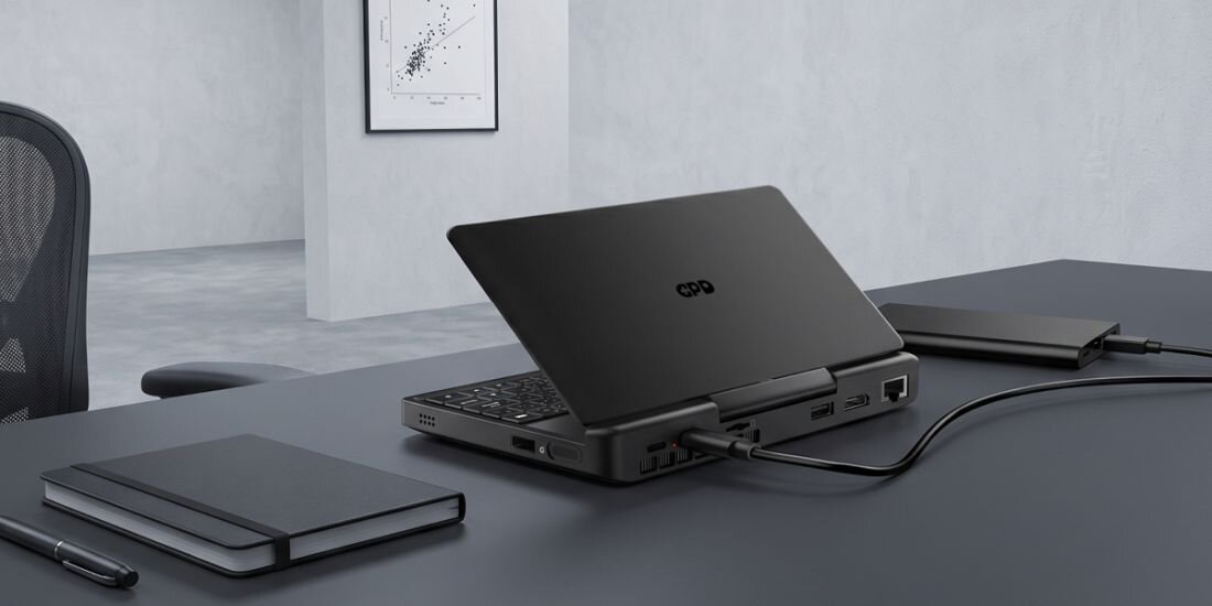 Na biurku znajduje się otwarty laptop z logo GPD, obok którego leży zamknięty notes i długopis. W tle widać krzesło biurowe i obraz na ścianie.