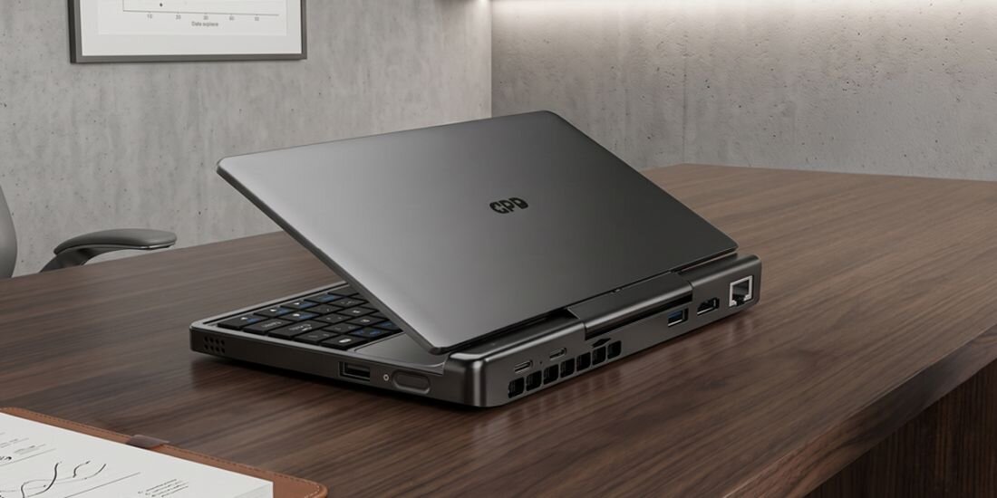 Na drewnianym biurku znajduje się mały, otwarty laptop z widocznym logo \'GPD\' na pokrywie. Laptop ma kilka portów po bokach, w tym USB i Ethernet.