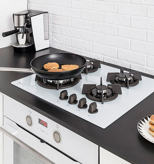 Na blacie kuchennym znajduje się biała płyta gazowa z czterema palnikami, na jednym z nich stoi patelnia z naleśnikami. Obok płyty widoczny jest ekspres do kawy, a poniżej piekarnik z wyświetlaczem pokazującym \'12:50\'.