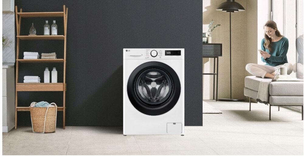 Biała pralka z dużym okrągłym bębnem i panelem sterowania z wyświetlaczem cyfrowym stoi na tle ciemnej ściany. Obok znajduje się drewniany regał z ręcznikami i butelkami, a w tle widać osobę siedzącą na sofie.