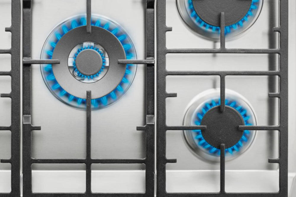 Obraz przedstawia płytę gazową z trzema palnikami, na których widać niebieskie płomienie. Palniki są otoczone metalowymi rusztami.