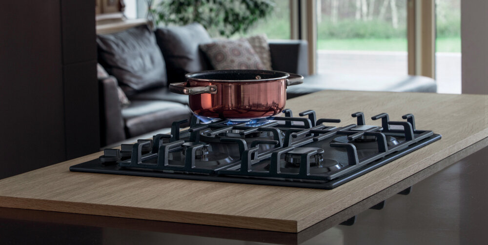 Na drewnianym blacie kuchennym znajduje się płyta gazowa z żeliwnymi rusztami, na której stoi czerwony garnek z niebieskim płomieniem pod spodem. W tle widoczna jest część salonu z sofą i dużym oknem.